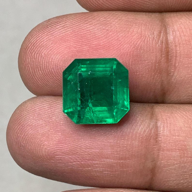 May include: A vibrant, emerald-cut green gemstone. The gem is held between fingers, showcasing its clarity and rich color. The cut features a rectangular shape with beveled corners, reflecting light and enhancing its brilliance. This gemstone is a beautiful example of fine jewelry.