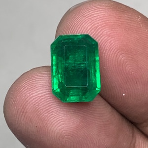 May include: A vibrant green emerald gemstone, cut in a rectangular shape with beveled edges. The emerald is set against a neutral background.