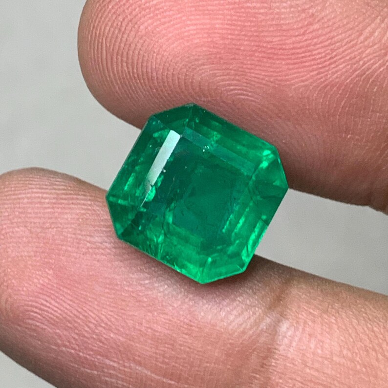 May include: A faceted, emerald-cut emerald gemstone. The gem is a vibrant green color and has a rectangular shape with beveled edges. The emerald is held between two fingers.