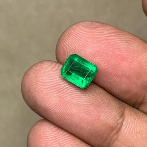May include: A vibrant, rectangular-cut emerald gemstone. The gem displays a rich, deep green color with visible internal inclusions. The emerald is held between fingers, showcasing its clarity and brilliance. This gemstone is a beautiful example of natural emeralds.