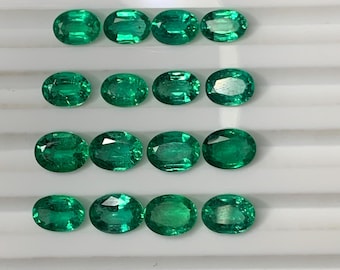 May include: A collection of 15 oval-shaped, green emeralds. The emeralds are arranged in five rows of three, each row on a separate white surface.