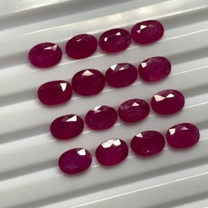 Könnte beinhalten: Eine Sammlung von 17 ovalen Rubin-Edelsteinen, die in Reihen auf einer weißen Oberfläche angeordnet sind. Die Rubine sind tiefrot und haben eine polierte, facettierte Oberfläche.
