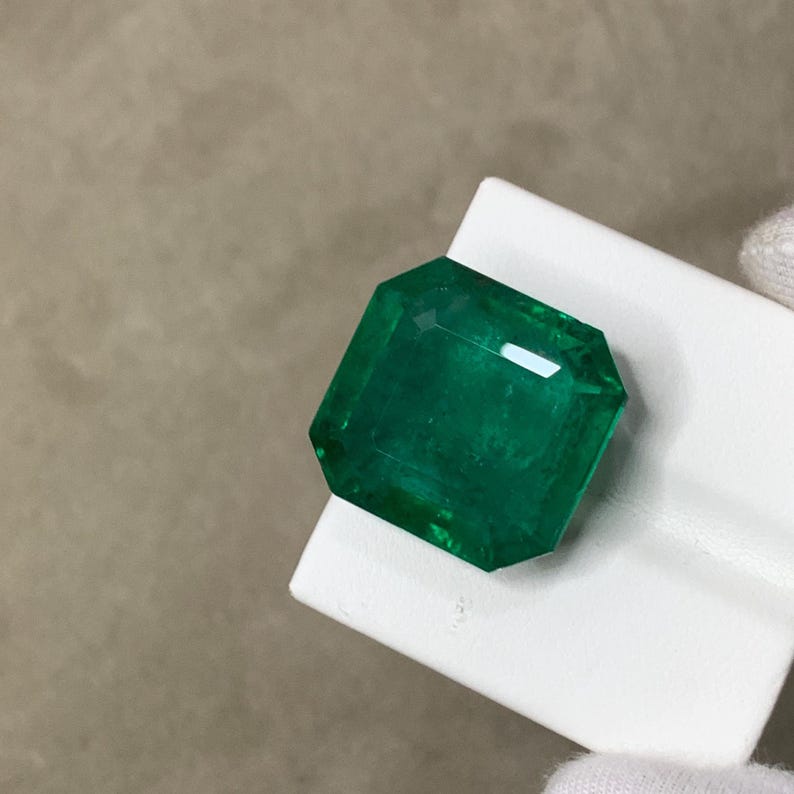 May include: A vibrant, emerald-cut green gemstone. The gem is displayed on a white surface, showcasing its facets and clarity. The stone's rich color and cut create a striking visual appeal, suggesting a high-quality precious stone.