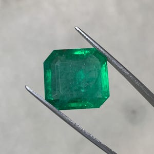 May include: A vibrant, emerald-cut emerald gemstone, held by a pair of tweezers. The gem displays a rich, deep green color and a faceted surface. The tweezers are silver-colored, and the background is a neutral gray.