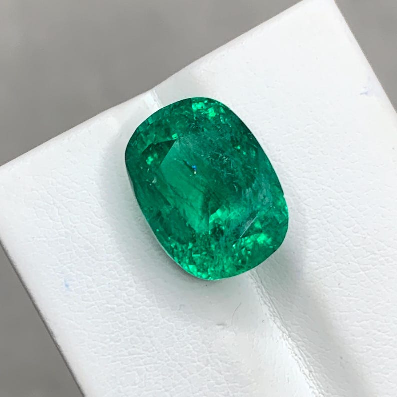 May include: A vibrant green emerald gemstone with a cushion cut. The emerald is set on a white background.