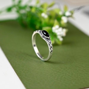 May include: A silver ring featuring an oval black gemstone. The ring's band and the area around the gemstone are decorated with an intricate pattern. The ring is set against a green backdrop, with white flowers visible.