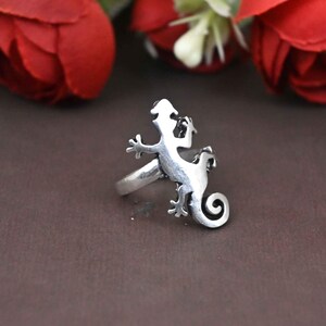 May include: A silver-colored ring featuring a detailed lizard design. The lizard has a curled tail and is positioned on a simple band. The ring is set against a brown background with red flowers in the background.