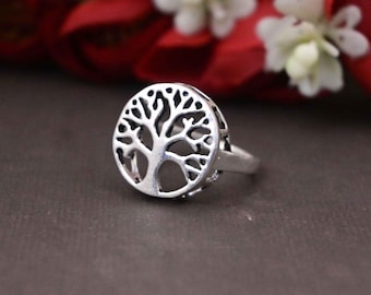 May include: A silver ring featuring a tree of life design. The ring has a circular pendant with a detailed tree silhouette. The ring is set against a dark background with red and white flowers.