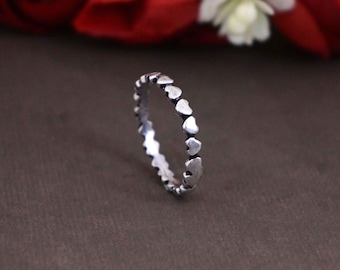May include: A silver ring featuring a repeating heart design. The ring is set against a dark brown background, with red and white flowers in the background. The ring is a delicate piece of jewelry.