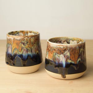 May include: Two ceramic tumblers with a unique glaze. The glaze features a blend of colors, including brown, blue, and gold, with a dark base and a light beige bottom. The tumblers are handmade and suitable for beverages.