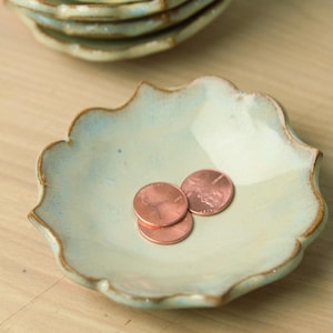 May include: A stack of small, light blue ceramic dishes with a scalloped edge and a brown rim. One dish in the foreground holds three copper pennies. The dishes are on a light wood surface.