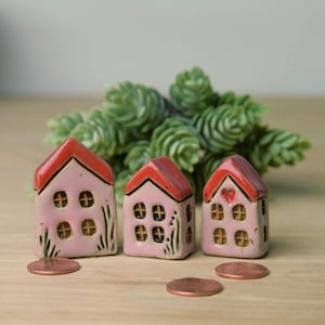 May include: Three miniature ceramic houses with pink walls and red roofs. Each house has four round windows and is decorated with small details. The houses are arranged on a wooden surface with a green plant in the background.