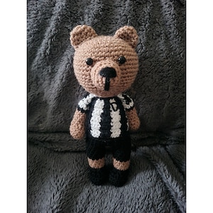 May include: A brown crocheted teddy bear wearing a black and white striped jersey. The bear has black eyes and a black nose.