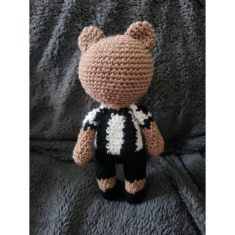 May include: A crocheted brown bear wearing a black and white striped shirt and black pants. The bear is standing on a gray fuzzy surface.