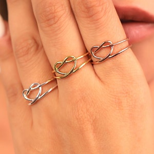 May include: Three thin metal rings worn on three fingers, each featuring a delicate open heart-shaped design. The rings are finished in three distinct metallic tones: a silver-colored ring, a gold-colored ring, and a rose gold-colored ring.