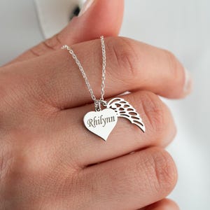 May include: A silver-toned necklace featuring a heart-shaped pendant engraved with the name Rhilynn and a separate wing-shaped charm. The charms are suspended from a delicate silver-toned chain, creating a layered look with two distinct metal pieces.