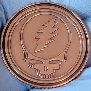 May include: A copper-colored round coin featuring a skull with a lightning bolt on its forehead. The coin has a detailed, ridged edge and a circular design. The skull has a smiling mouth with visible teeth.