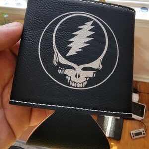 May include: Black beverage holder with a white stitched edge and a white graphic of a skull with a lightning bolt on the front. The holder is made of a textured material.