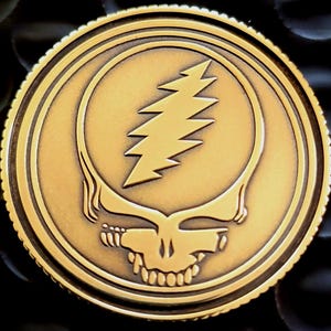 May include: A gold-colored coin featuring the Steal Your Face logo, a skull with a lightning bolt. The coin has a detailed, embossed design with a ridged edge. The background is dark.