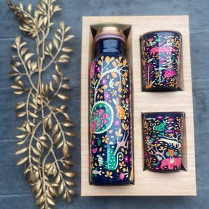 May include: A navy blue water bottle and two matching glasses with a colourful floral and animal design. The bottle and glasses are in a wooden box. The bottle has a copper-coloured lid. A gold-coloured decorative branch is on the left.