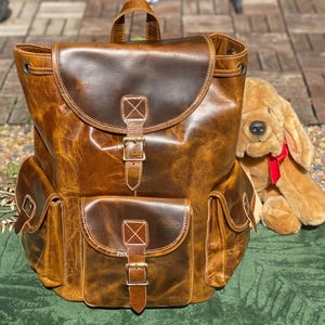 May include: A brown leather backpack with two large front pockets and a buckle closure. The backpack has a distressed look and is made of high-quality leather.