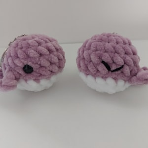 May include: Two handmade, plush whale keyrings. The whales are lavender with white bellies and small black eyes. The keyrings are made of a soft, textured material.