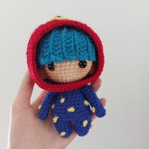 May include: A handmade crochet doll with a blue knitted cap and a red hood. The doll wears a blue jumpsuit with yellow polka dots. The doll's face is tan with black eyes. The doll is held in a hand.