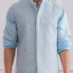 Peut inclure: Un gilet brodé bleu clair avec un col mandarin, porté sur une chemise à manches longues assortie. Le gilet présente un motif détaillé et répétitif. Les manches de la chemise sont retroussées.