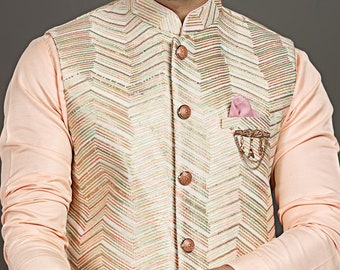 May include: A cream-colored, chevron-patterned vest with brown buttons and a pink pocket square. The vest is worn over a light peach-colored long-sleeved shirt. A gold ring is visible on the person's finger. The vest has a decorative chain.