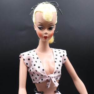 May include: A vintage doll with blonde hair, wearing a pink and black polka dot swimsuit top. The doll has a painted face with red lips and black eyes.