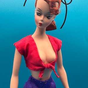 May include: A vintage doll with red hair styled in a side part and a black hair accessory. She wears a red tie-front top and purple trousers. The doll has painted facial features, including red lipstick and dark eye makeup.