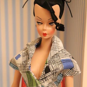 May include: A vintage doll with black hair styled in a ponytail with a black ribbon. She is wearing a white and black checked shirt with blue accents and a turquoise skirt.
