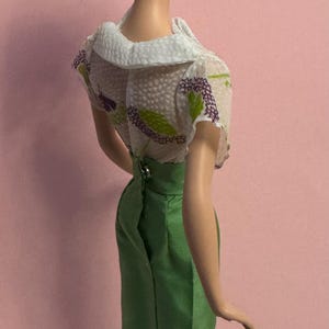 May include: A vintage doll in a white floral blouse and a green skirt. The doll has black hair in a ponytail, black earrings and shoes. The doll stands on a black and white chequered surface. The doll's height is approximately 30 cm.