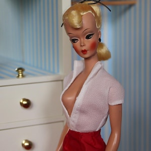 May include: A vintage doll with blonde hair styled in a bouffant, wearing a white collared shirt with a low neckline and a red skirt. The doll has a painted face with red lips and black eyes.