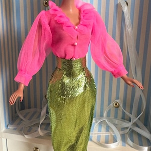 May include: A vintage doll dressed in a bright pink blouse with a ruffled collar and long, sheer sleeves. The doll is also wearing a shimmering, lime green pencil skirt. The doll has blonde hair and is standing on a clear stand.
