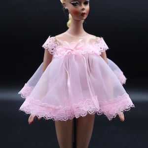 May include: A vintage doll wearing a pink lace nightgown with a bow at the neckline. The doll has blonde hair and is wearing black earrings.