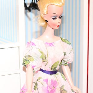 May include: A doll with blonde hair wearing a pink dress with a floral pattern and a purple belt. The doll is standing in a dollhouse with blue and white striped wallpaper.