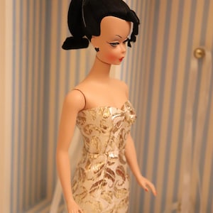 May include: A vintage doll with black hair styled in a ponytail, wearing a gold and white patterned strapless dress. The doll is standing in front of a striped background.