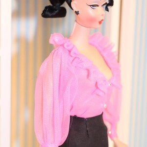 May include: A doll with black hair styled in a ponytail with a black headband. She is wearing a pink ruffled blouse and a black skirt.