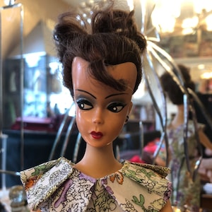 May include: A vintage doll with brown hair styled in a bun, wearing a white dress with a colourful floral print and a large collar.