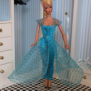 May include: A vintage Barbie doll wearing a blue jumpsuit with a matching sheer overlay featuring a polka dot pattern. The doll has blonde hair and is wearing black heels.