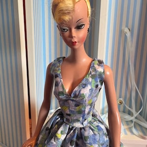 May include: A vintage doll with blonde hair in a bun, wearing a blue floral print dress. The doll has painted facial features, including dark eyeliner and red lipstick. The dress has a V-neck and a full skirt.