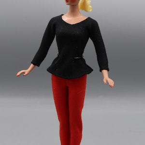 May include: A vintage doll with blonde hair styled in a ponytail, wearing a black long-sleeved shirt with a black belt and red trousers. The doll is standing on a black stand.