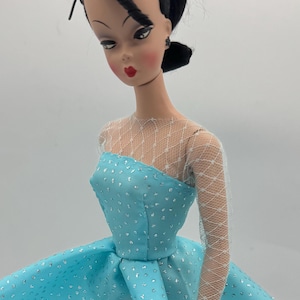 May include: A vintage doll with black hair and a blue dress with silver glitter. The doll is wearing a white lace top and has red lips.