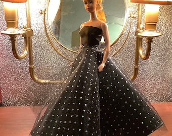 May include: A black and silver sequined gown with a fitted bodice and a full skirt. The gown is worn by a fashion doll with blonde hair and a black headband.