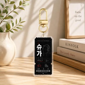Peut inclure: Un porte-clés en acrylique transparent en forme de billet de concert avec un fermoir en métal doré. Le billet présente un fond noir avec du texte blanc, incluant les caractères coréens pour Suga, le nom Agust D et un chat portant un casque.