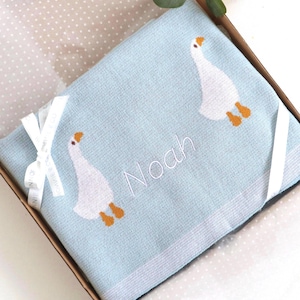 May include: A light blue baby blanket with embroidered white geese and the name "Noah" in white script. The blanket is folded in a gift box and tied with a white ribbon.