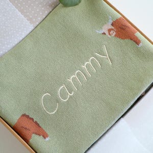 May include: A light green blanket with the name "Cammy" embroidered in white script. The blanket features brown and white Highland cow illustrations in the corners. The blanket is folded and presented in a light brown box.