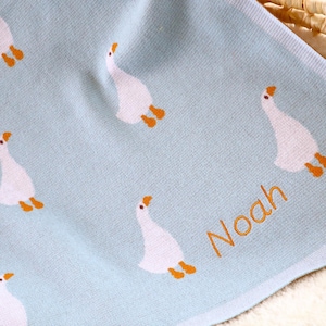 May include: A light blue baby blanket featuring a pattern of white geese with orange feet and beaks. The name "Noah" is embroidered in orange thread on the blanket. The blanket is made of a soft, textured material.