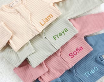 May include: A collection of ribbed baby zip-up cardigans in pastel colors: cream, sage green, pink, and blue. Each cardigan has a name embroidered on the front in a contrasting color: Liam, Freya, Sofia, and Theo. The cardigans are arranged on a soft, white surface.